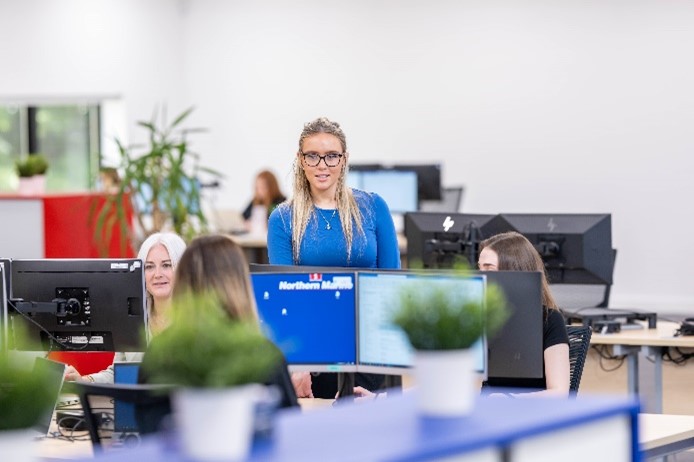 An office scene with a woman in a blue top standing and talking to colleagues seated at desks with computer monitors.