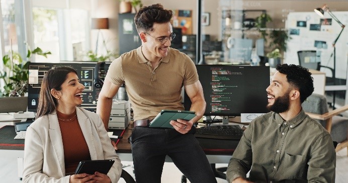 Three colleagues in a modern office smiling and discussing, with computer screens displaying code in the background.