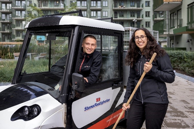 Two workers in a residential area, one in a utility vehicle and the other holding a broom, both wearing Stena Fastigheter uniforms.