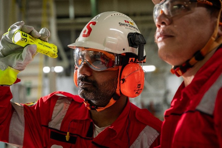 Two workers in red uniforms and safety gear inspect equipment in an industrial setting, with one holding a yellow tool.