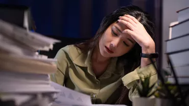 Stressed woman in office, facing work stress and burnout, surrounded by documents at her desk.