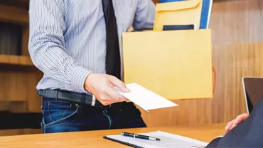  An employee, wearing a shirt and tie, extends a resignation letter to their boss while holding a box of documents.