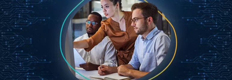 Two colleagues sit at a desk and take notes, while a woman standing above them directs their attention towards a screen.